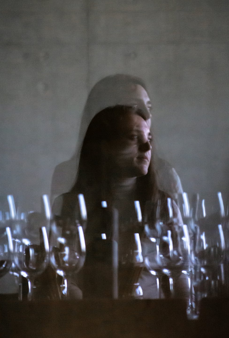 Woman with dark hair looks thoughtfully past blurred wine glasses.
